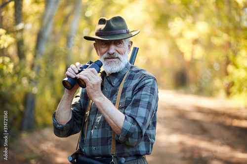 Using binoculars for better hunting on birds. Senior man holding binoculars during hunting in autumn forest look at camera. Road background