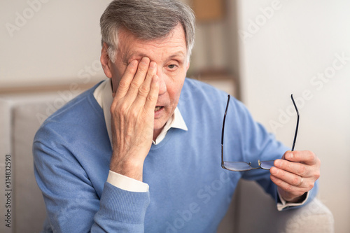 Tired Elderly Man Massaging Eye Sitting On Couch At Home