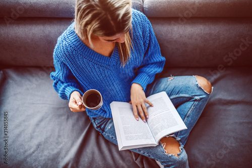 Top view of blond caucasian young woman in sweater sitting on sofa in living room, drinking coffee and reading book.