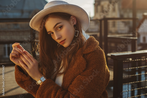 Outdoor fashion portrait of young elegant fashionable brunette woman, model wearing stylish white hat, wrist watch, brown faux fur coat, posing at sunset, in European city. Copy empty space for text