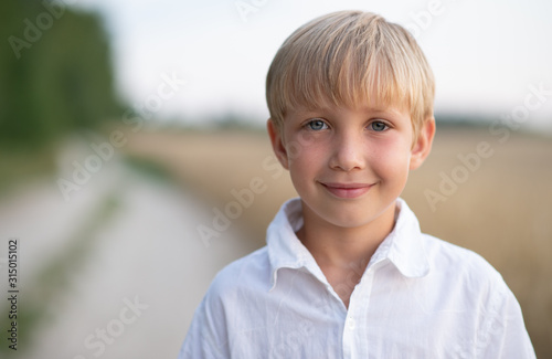 Face close - up of a boy of eight years. Portrait of a smiling boy in nature looking at the camera. Happy blond boy with a smile on his face. Child in a white shirt with blue eyes in nature.