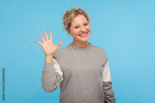 Hello! Cheerful woman with short hair in casual sweatshirt smiling friendly at camera and waving hi, welcoming or showing goodbye, hospitable greeting. indoor studio shot isolated on blue background