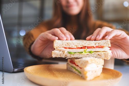 Closeup image of a woman holding and eating whole wheat sandwich while working on laptop computer