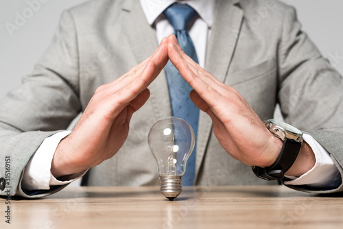 cropped view of businessman showing protection gesture above light bulb isolated on grey