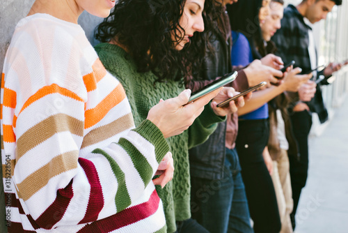 Young men and women look at their smart phones - Standing against a wall in the City - Concept of tech obsession