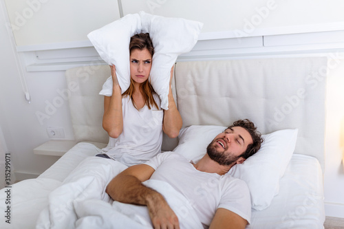 Young couple have problem with man's snoring. Heterosexual couple in bed, man sleeps and snoring with mouth open, while a tired woman irritated by snoring sitting on bed with a pillow on her head.