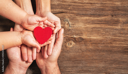 family hands holding red heart on wooden background. Donation, charity, health concept.