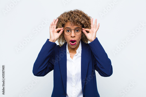 young woman african american feeling shocked, amazed and surprised, holding glasses with astonished, disbelieving look against flat wall