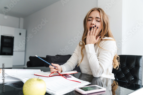 Photo of young sleepy woman yawning and making notes in exercise book