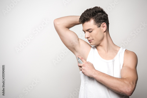 young man in white sleeveless shirt applying deodorant on underarm isolated on grey