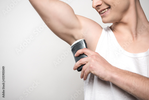 cropped view of smiling man applying deodorant on underarm isolated on grey