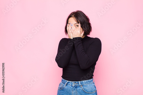 young pretty woman feeling scared or embarrassed, peeking or spying with eyes half-covered with hands against pink wall