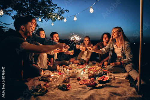 Happy friends having fun with fire sparkles. Young people millennials camping at picnic after sunset.