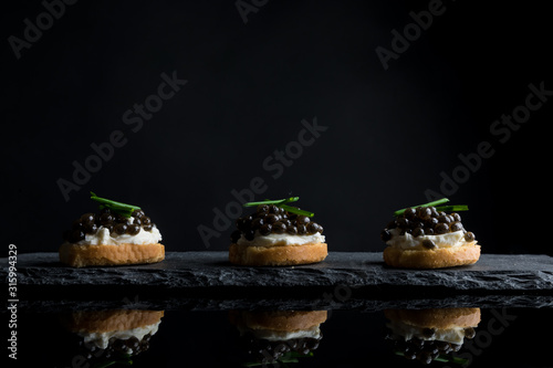 Caviar, three snacks of black caviar on bread and stone plate isolated on black background with reflections, copy space