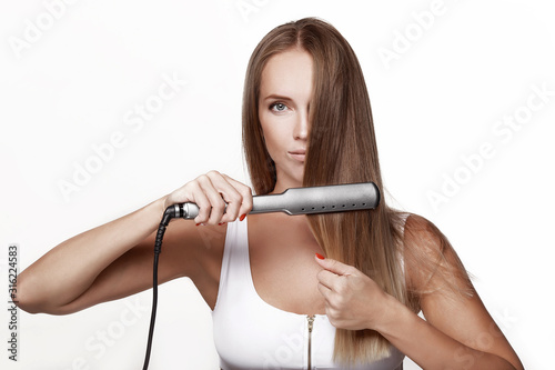 A beautiful young girl in a white blouse straightens her long hair with a hair straightening iron. Makes herself an evening hairstyle, a beauty salon. Covers one eye with hair. On white background.