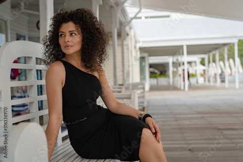 A beautiful young curly-hair woman in a little black dress is posing on a bench. Portrait Photography. 