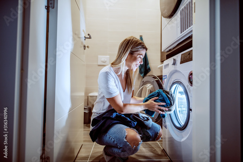 Smiling caucasian blond worthy housewife dressed casual crouching in bathroom and loading washing machine with dirty clothes in late hours.