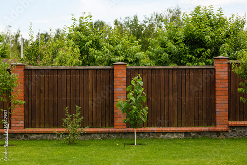 New wooden fence with massive stone brick pillars. Green lawn and trees, daytime