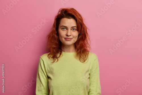 Portrait of attractive young female has natural red hair, looks directly at camera with gentle smile, wears green casual jumper, isolated over pink background. Human facial expressions concept