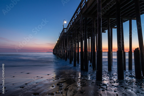 Ventura Pier at sunset, Ventura, California. Rocks and sand in foreground, water receded in low tide. Lamps on pier; colored twilight sky in background.