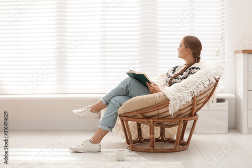 Young woman reading book in papasan chair near window at home