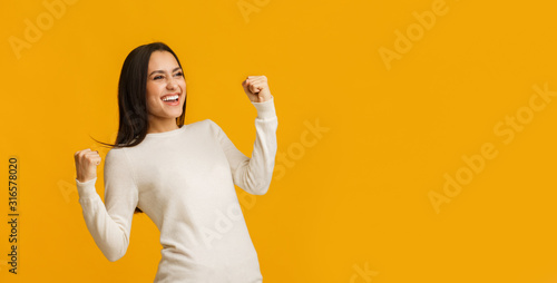 Euphoric brunette woman celebrating success with raised fists over yellow background