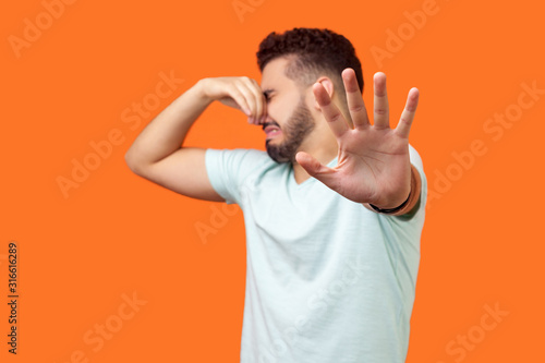 Stinky smell. Side view of confused brunette man with beard in white t-shirt holding breath with fingers on nose, disgusted by bad odor fart, gesturing stop. studio shot isolated on orange background