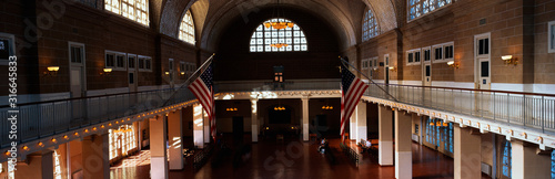 This is the interior of the Great Hall at Ellis Island which signifies immigration to the United States. There are two American flags flying posted at the center of the walls.