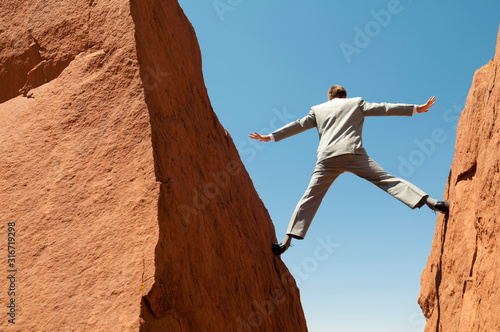 Unrecognizable businessman balancing outdoors between two jagged rocks forming a deep crevasse against blue sky