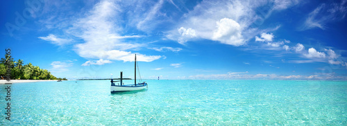 Boat in turquoise ocean water against blue sky with white clouds and tropical island. Natural landscape for summer vacation, panoramic view.