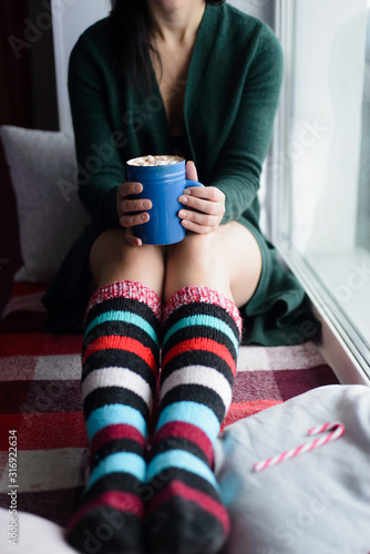 white caucasian woman in green knitted cardigan and colorful socks holding blue cup with cacao and marshmallow, front view, vertical stock photo image