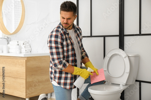 Young man feeling disgust while cleaning toilet bowl in bathroom