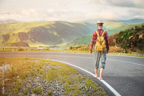 Stylish barefoot bearded male hitchhiker traveler in a hat and with a backpack walks along a country road in the mountains at sunset. The view from the back. Travel concept without money.