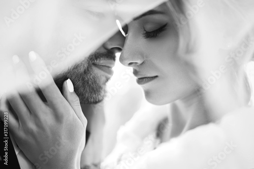 Black and white foto of amazing smiling wedding couple. Pretty bride and stylish groom posing and kisses tenderly in the shadow of a flying veil. Romantic moment.Together. Wedding. Marriage.