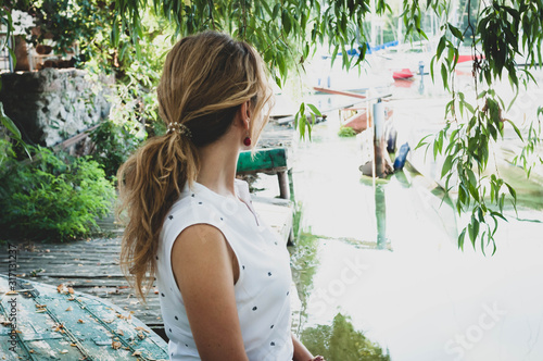 side view of woman in white blouse with ponytail looking at boats and sitting in front of tree branches near river