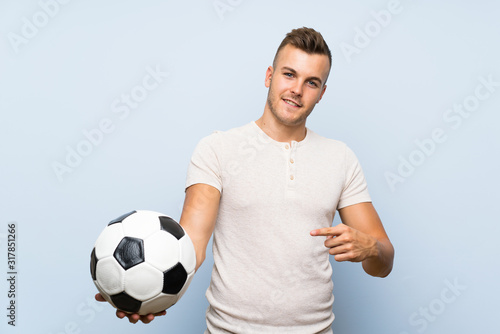 Young handsome blonde man over isolated blue background holding a soccer ball