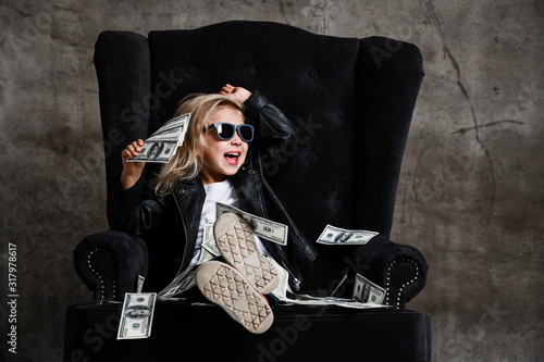 Happy screaming loud laughing kid girl is sitting in big plush armchair holding a wad of cash dollars, throwing money