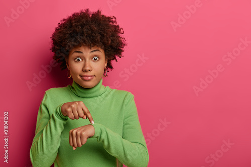 Time to go, hurry up! Shocked ethnic woman points at wrist, makes time gesture, shows we should do everything quickly, dressed in green clothes, stands against pink background. Its too late.