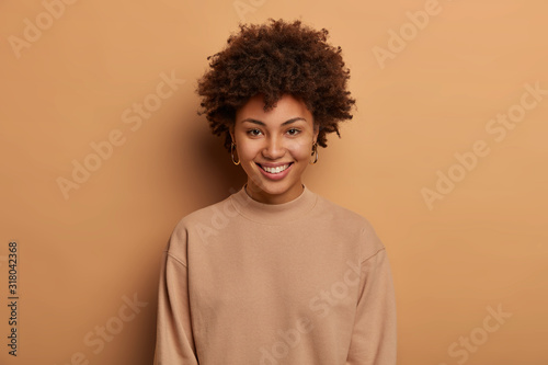 Modest relaxed healthy Afro American woman has tender toothy smile, enjoys lovely day, expresses positive emotions, wears casual brown jumper, looks directly at camera with eyes full of happiness.