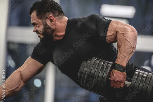 strong young bearded caucasian man in black sportswear lifting heavy weight dumbbell with one muscle with vein hand training back and arms in sport gym training routine