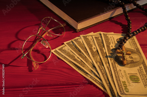 Rosary beads, money, glasses and holy bible. Church donation or tithe concept. Red wood table surface background.