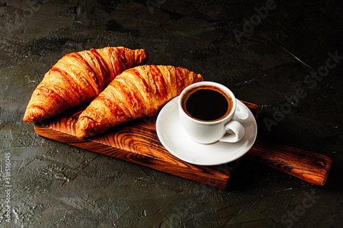 Appetizing breakfast, croissants and coffee in white cup with a spoon and a saucer on a wooden plate, on a dark gray table under concrete