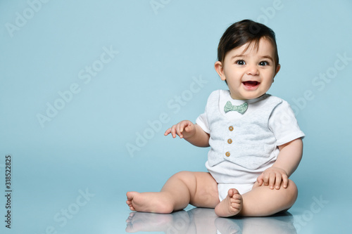 Little kid in white bodysuit as vest with bow-tie, barefoot. He smiling, sitting on the floor against blue background. Close up