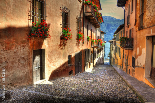 Narrow Street with Sunlight and Old Houses with Red Flowers and Lake with Mountain in Cannobio, Italy.