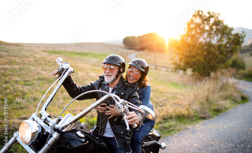 Cheerful senior couple travellers with motorbike in countryside at sunset.