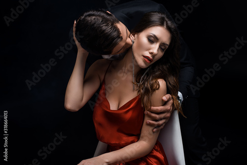 Elegant man kissing beautiful woman in red dress isolated on black
