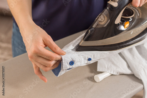 Kobiece dłonie prasują żelazkiem rękaw koszuli na podstawce stojącej na desce do prasowania. Wyprasowane ubrania leżą na stole w tle.