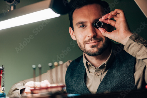 Selective focus of watchmaker holding eyeglass loupe and looking at camera by working table