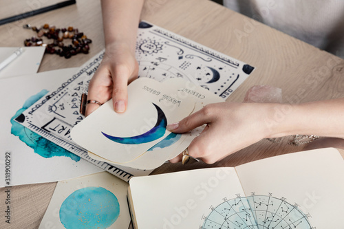 Cropped view of astrologer holding cards with watercolor drawings by birth chart with zodiac sign on table