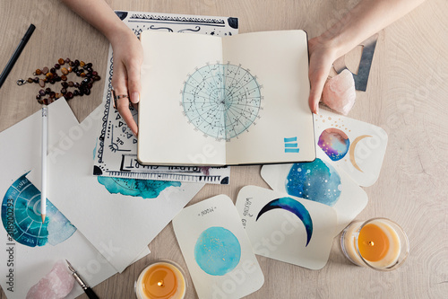 Top view of astrologer holding notebook with watercolor drawings and zodiac signs on cards on table